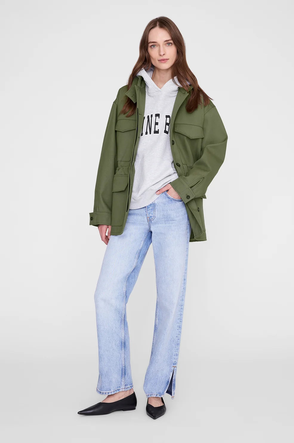 A woman modeling an olive green utility jacket over a white graphic hoodie, paired with light-wash blue jeans and black flats, against a plain light gray studio background.