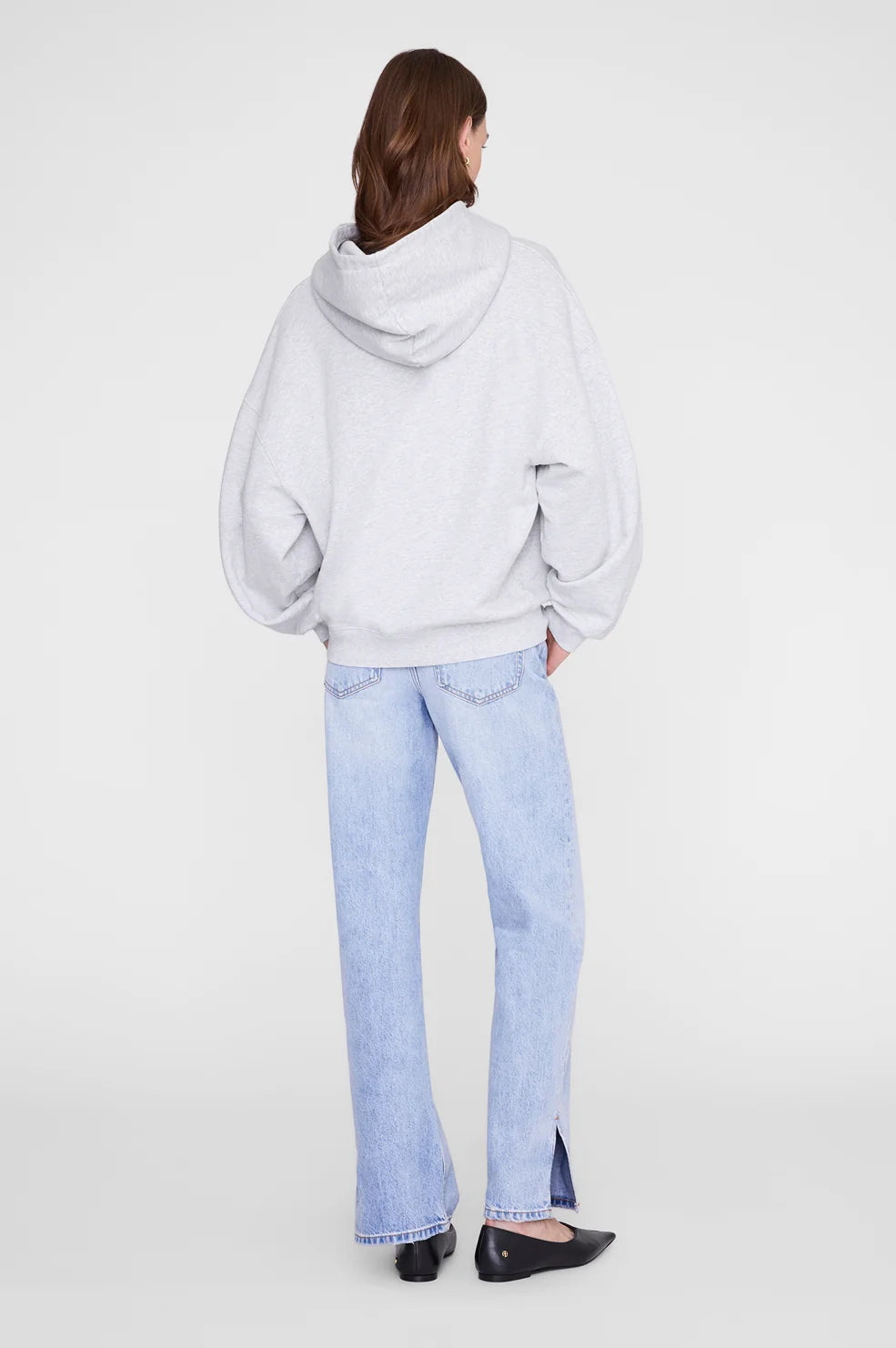 A model shown from the back wearing a light gray oversized hoodie, light-wash blue jeans, and black flats against a plain light gray studio background.