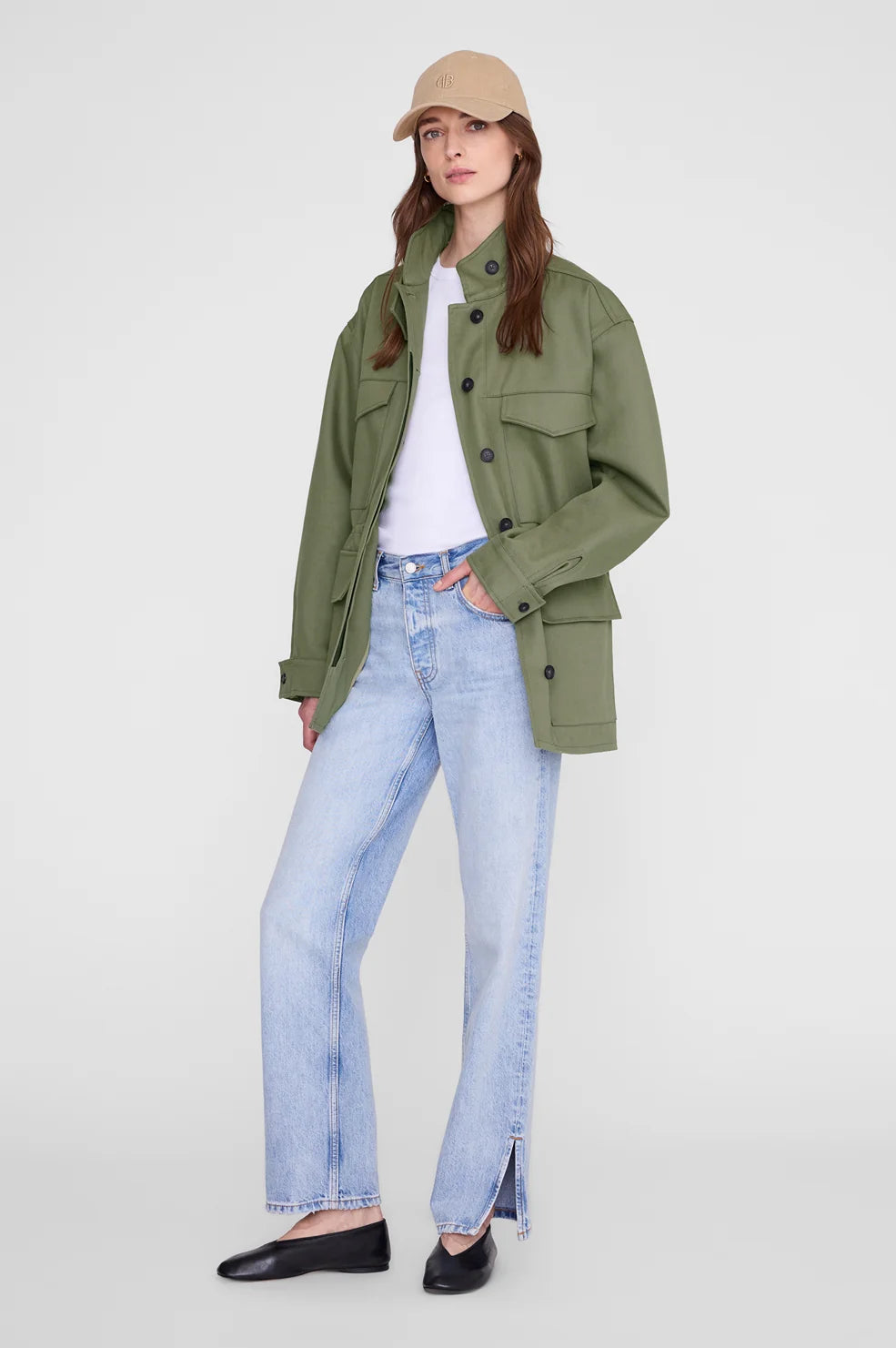 A model wearing an olive green utility jacket over a white t-shirt, light-wash blue jeans, and black flats, standing against a light gray studio background.