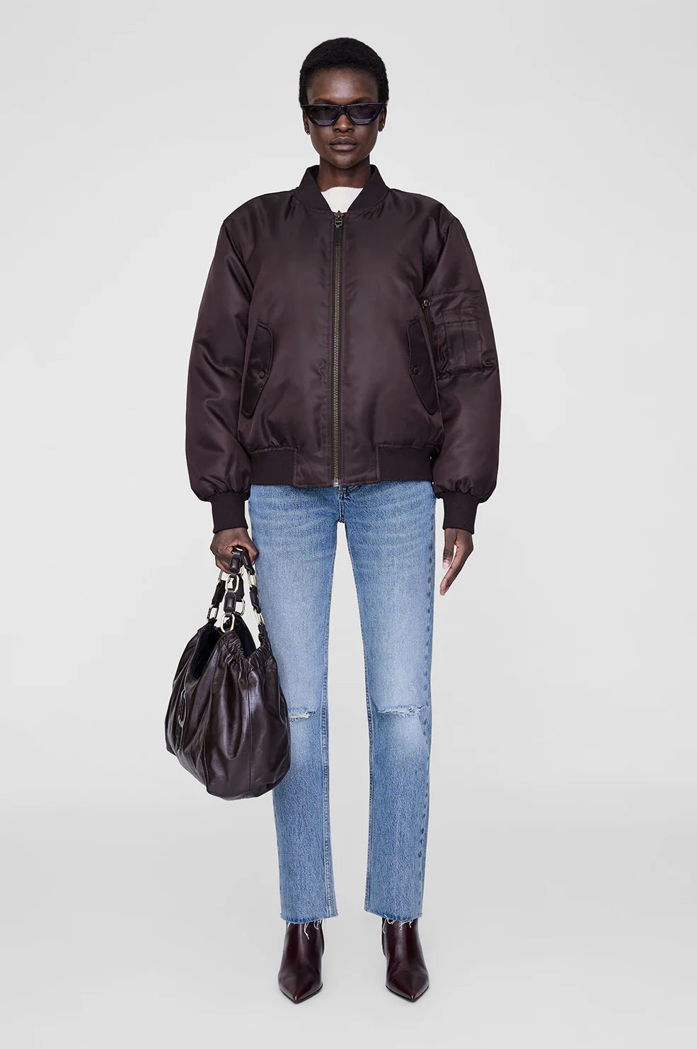 Person wearing a brown bomber jacket from Anine Bing and light blue jeans, holding a brown leather bag on a white background