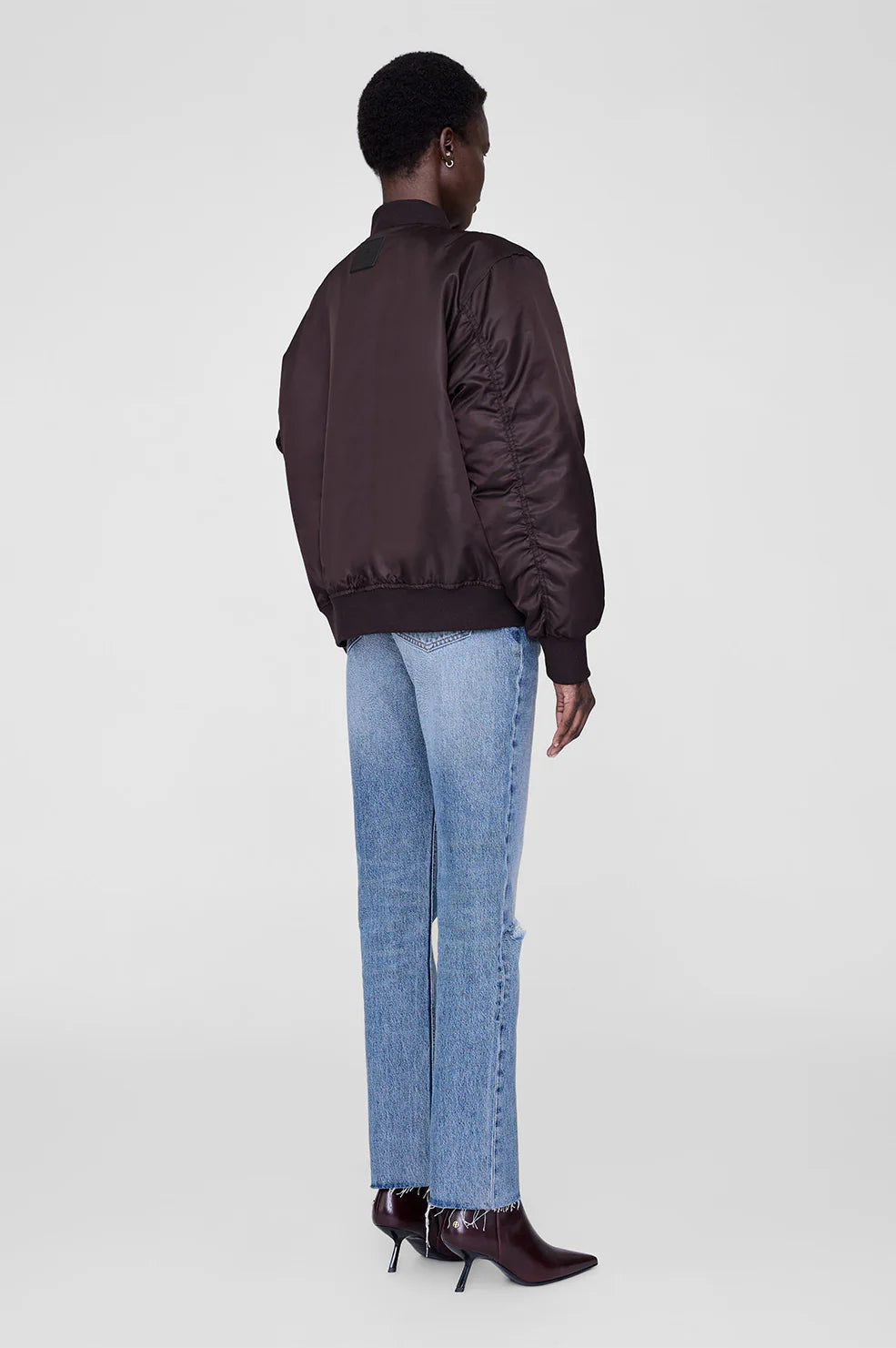Person wearing a dark brown bomber jacket and light blue jeans from Anine Bing on a white background