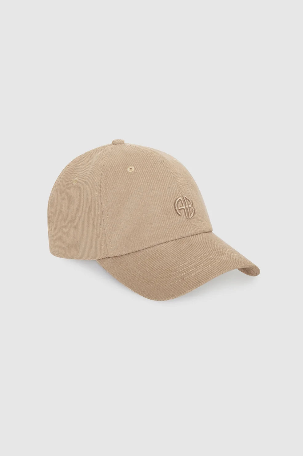 A beige baseball cap made of textured corduroy fabric, featuring a small embroidered circular monogram logo on the side, set against a solid light gray background.