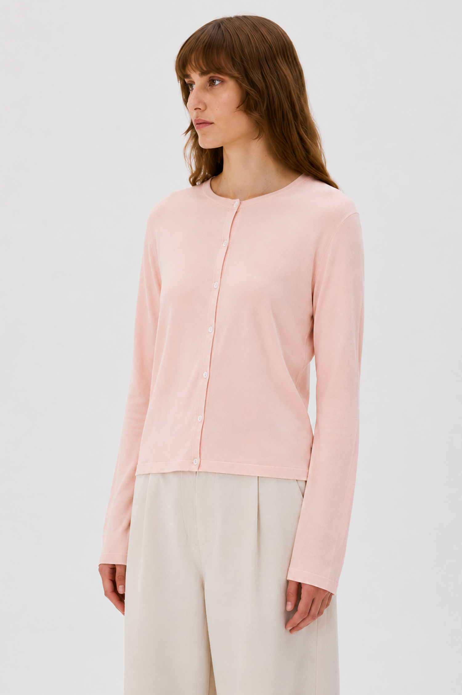 A woman wearing a light pink long-sleeve button-front cardigan and cream wide-leg trousers, photographed against a clean light gray studio background.