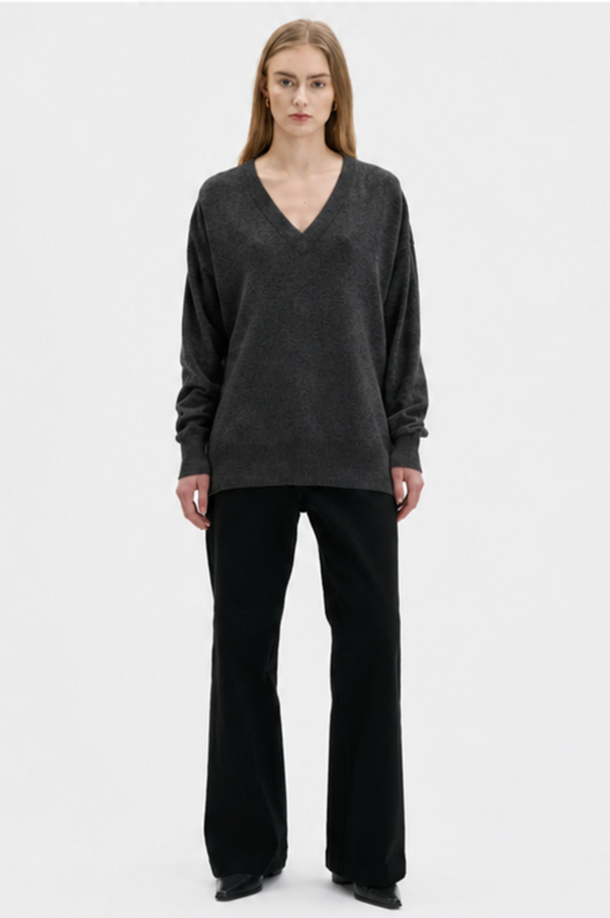 A model stands against a plain light gray studio background wearing a dark charcoal gray V-neck cashmere knit sweater and black wide-leg trousers.