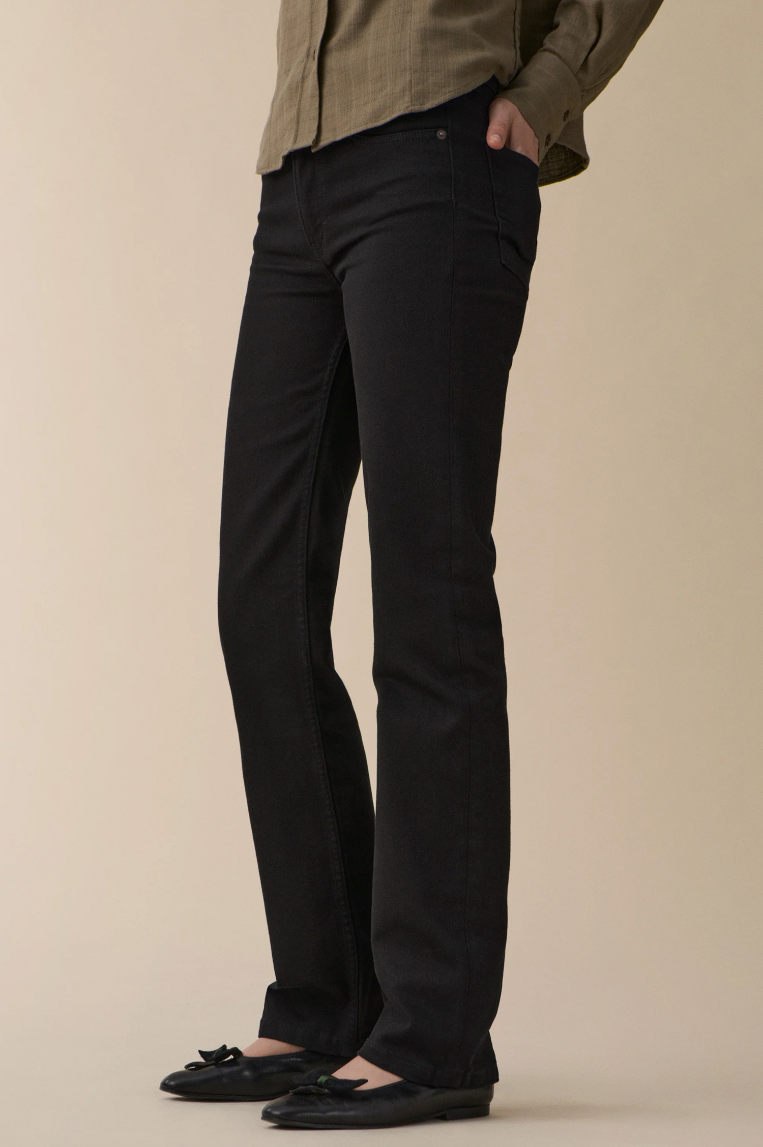 A person wearing black straight-leg or bootcut pants with a relaxed fit from jeanerica, paired with a muted olive green shirt and black flat shoes, standing against a solid beige studio background.