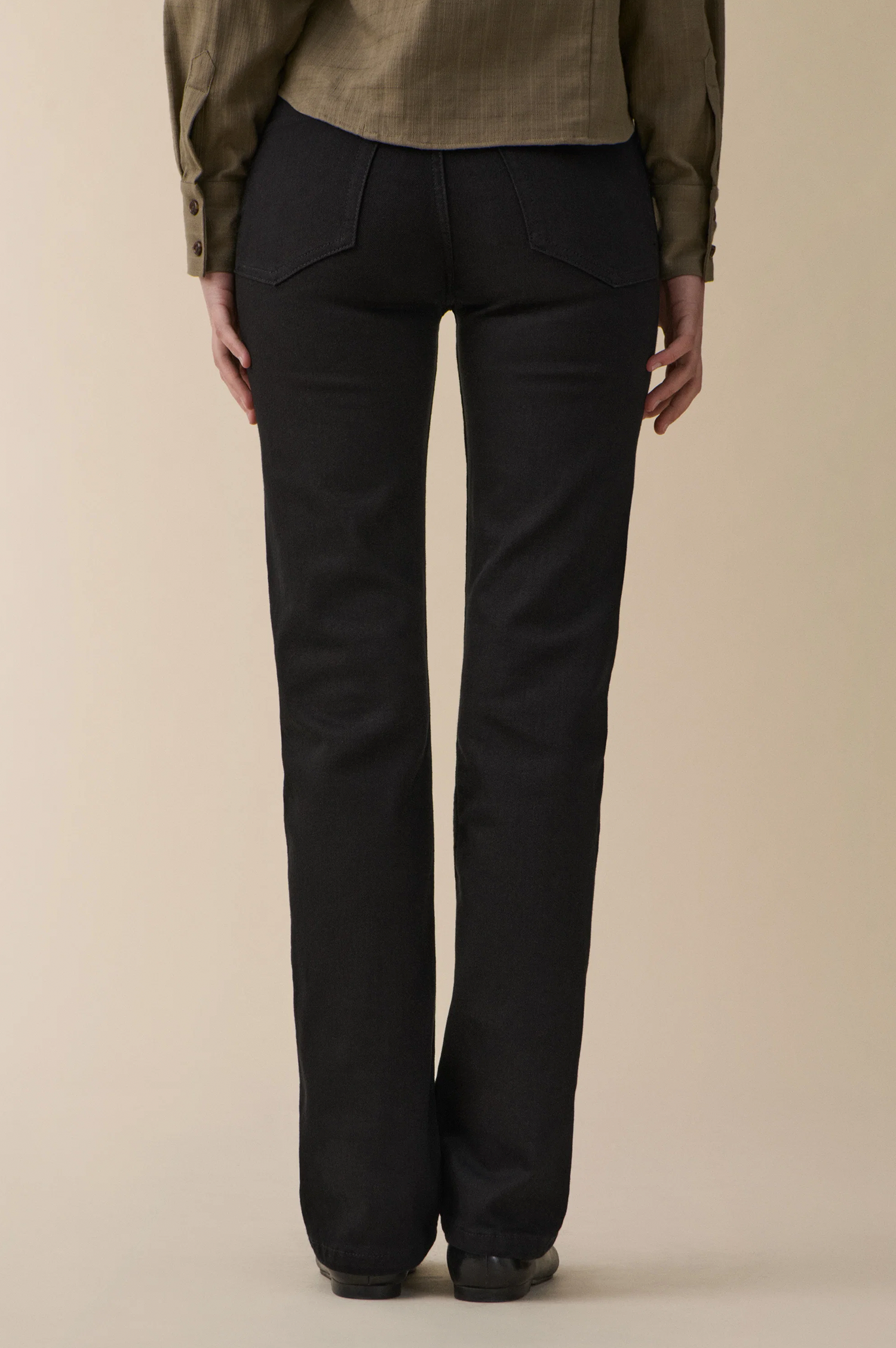 Rear view of a person wearing black straight-leg trousers with a relaxed fit and back pockets, from jenerica paired with an olive green shirt and black shoes, against a solid beige background.