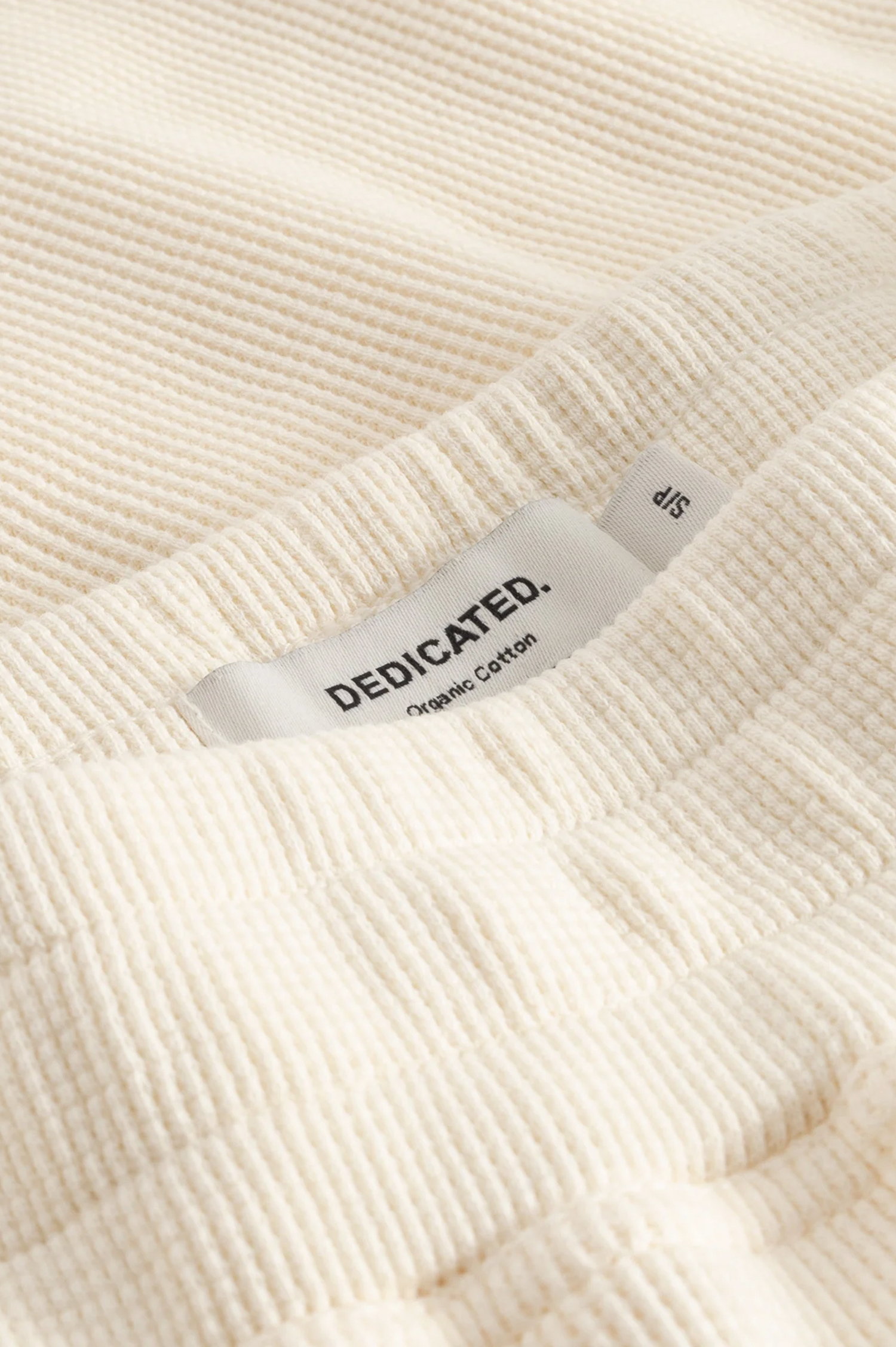 Close-up of a cream-colored ribbed knit garment with a visible fabric label reading 'DEDICATED. Organic Cotton' and size '4S'.