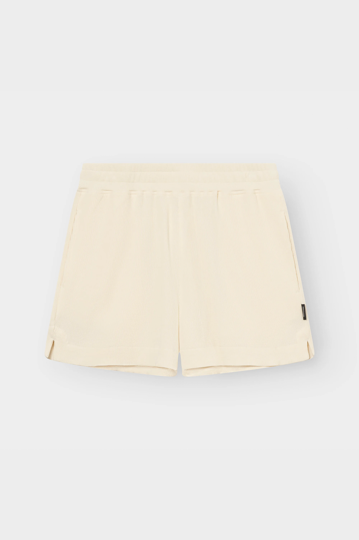 A pair of light cream or off-white athletic shorts with an elastic waistband, laid flat against a light gray background. A small black brand tag is visible on the lower left hem.