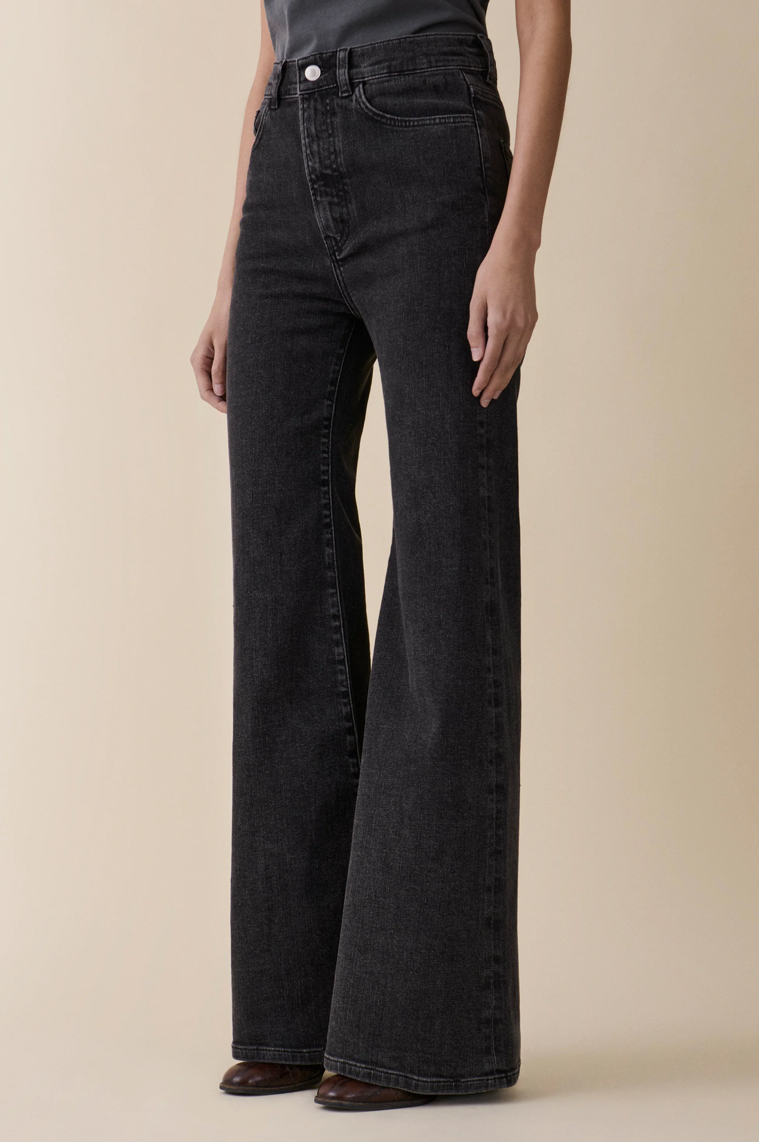 A model wearing a pair of Fuji wide-leg black denim jeans from jeanerica with a faded charcoal wash, paired with a dark gray top and brown leather shoes, set against a solid beige background.