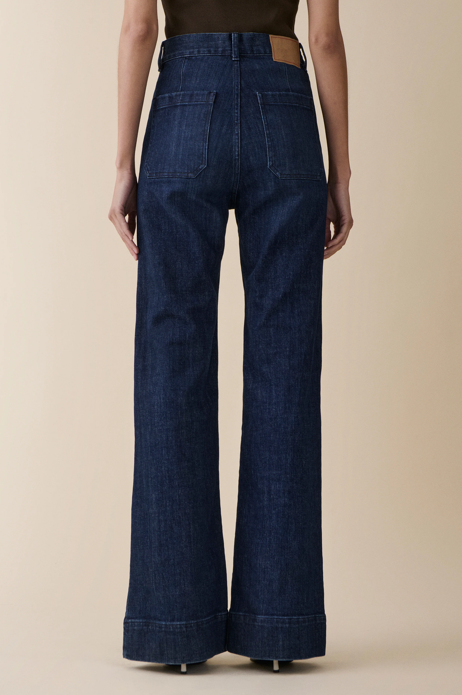 Rear view of a model wearing dark blue wide-leg denim jeans with a brown leather waistband patch, set against a solid beige background.