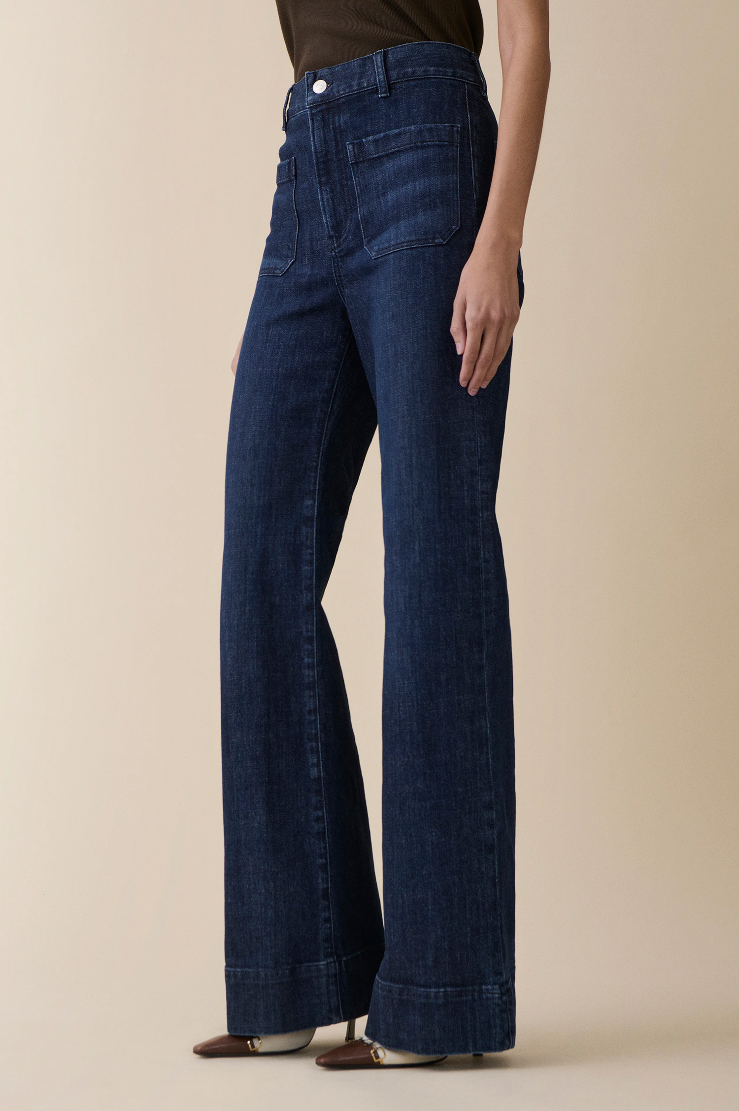 A model wearing dark blue high-waisted wide-leg jeans with rolled cuffs from jeanerica, paired with a dark brown top and brown pointed-toe flats, set against a solid beige studio background.