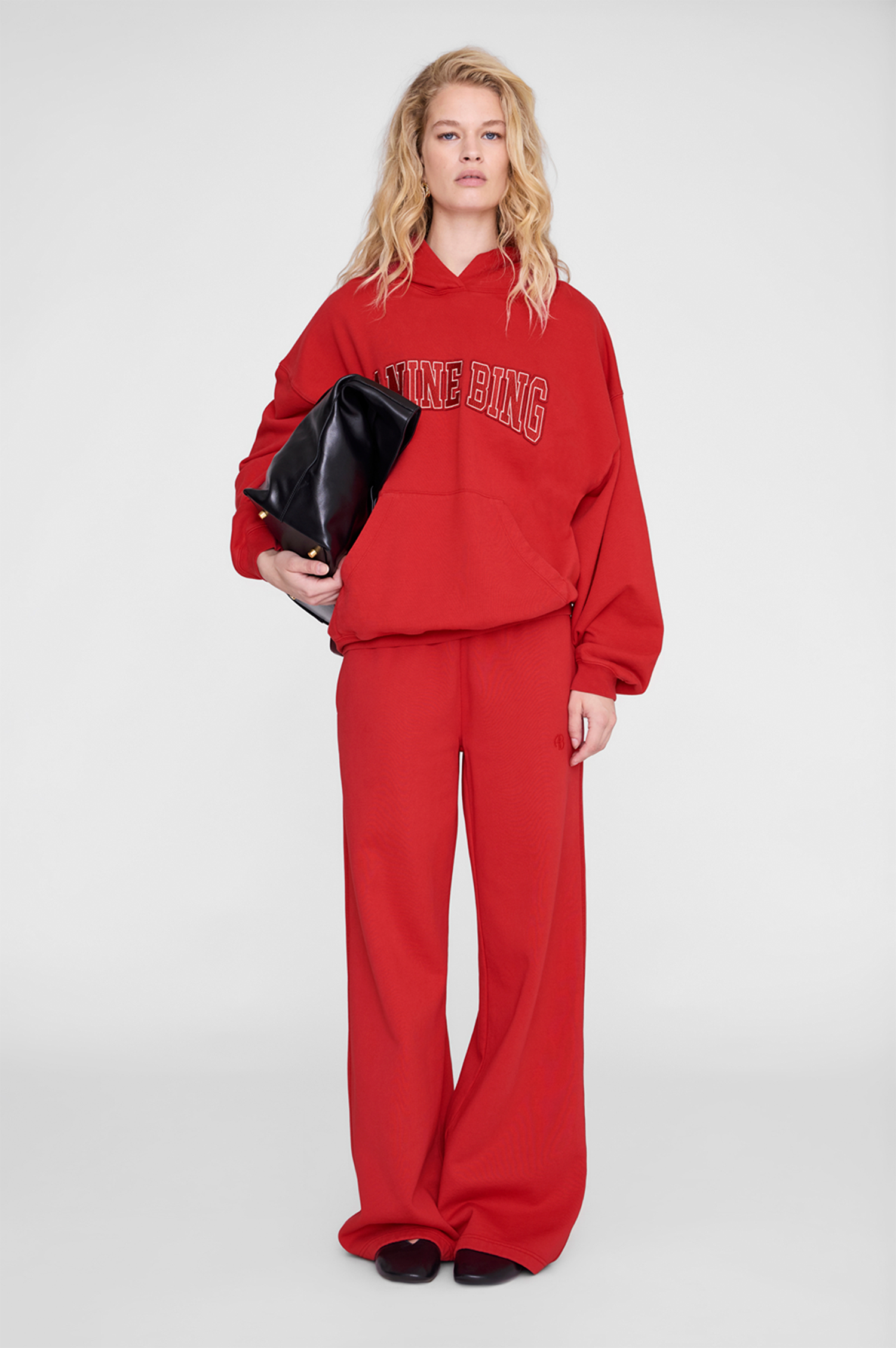 A model stands against a light gray studio background wearing a bright red two-piece lounge set consisting of a cropped hoodie with 'YONG BING' text across the chest and matching wide-leg pants. She is holding a black clutch bag.
