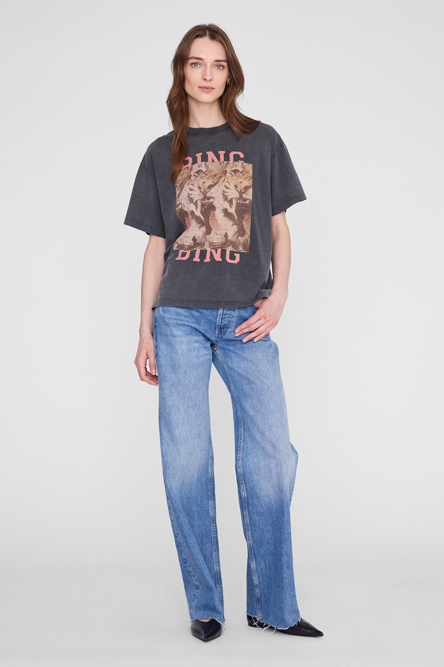 A woman modeling a dark gray graphic t-shirt with a vintage-style animal print and pink lettering, paired with wide-leg blue jeans, against a light gray studio background.