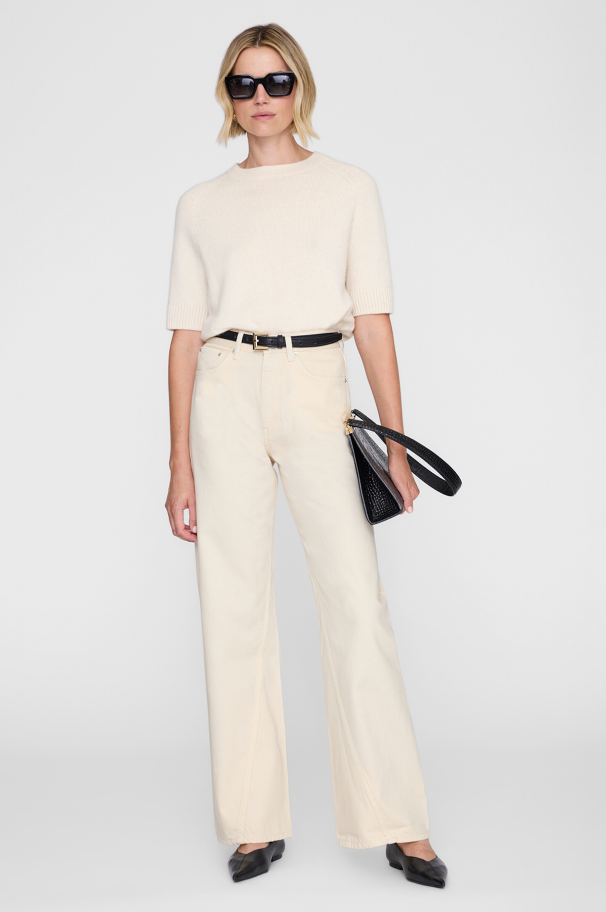Woman wearing a beige outfit with sunglasses and a black handbag on a white background