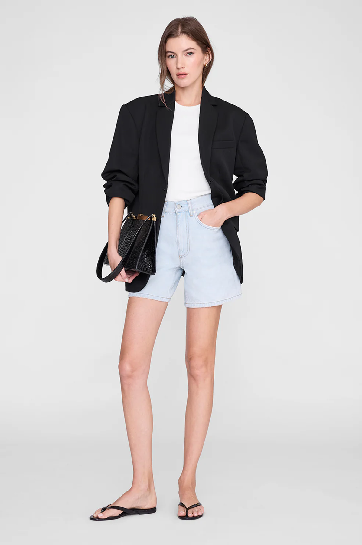 Woman wearing a black blazer, white top, light blue shorts, and black sandals on a white background