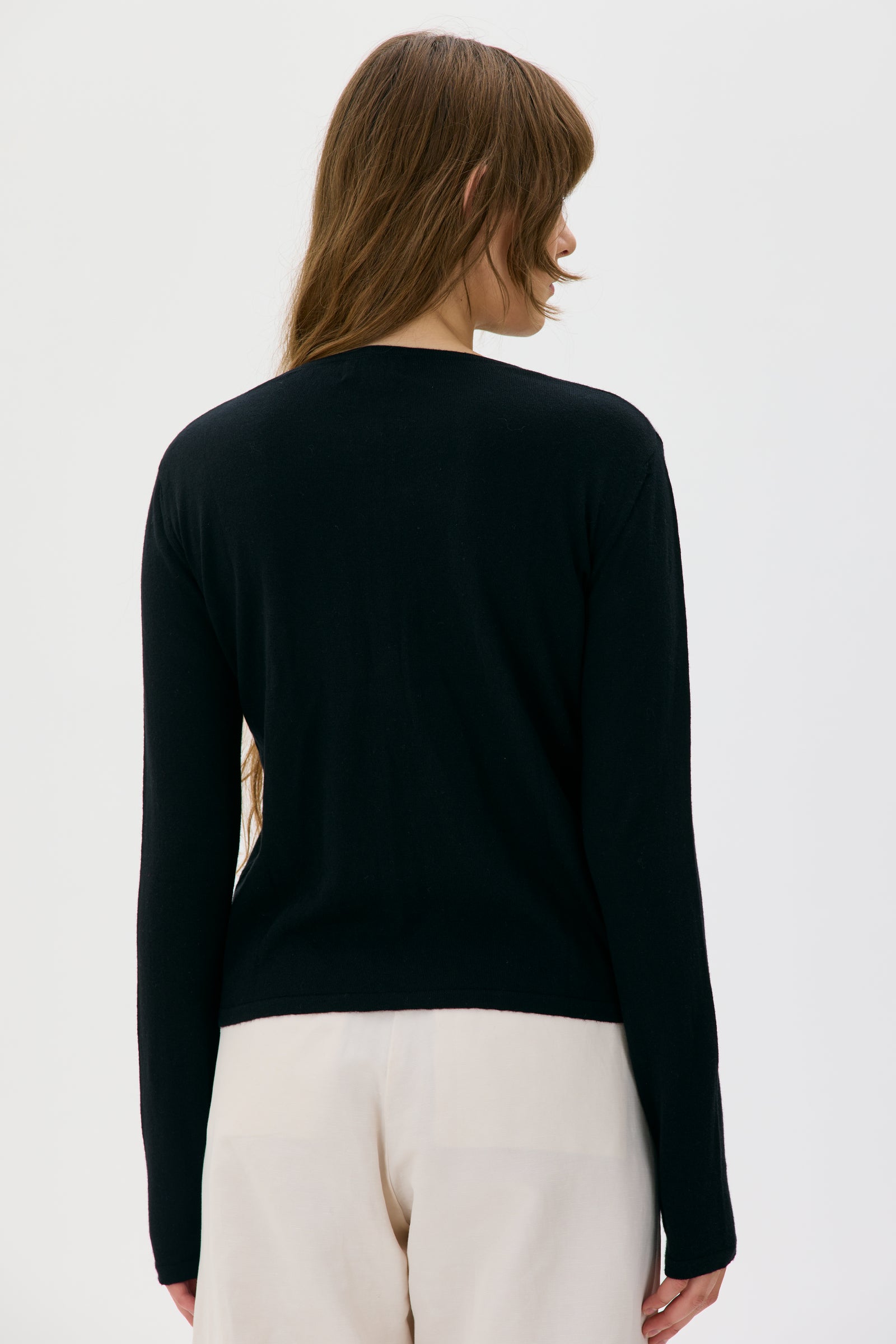Person wearing a black long-sleeve cardigan from maska and white pants on a light gray background