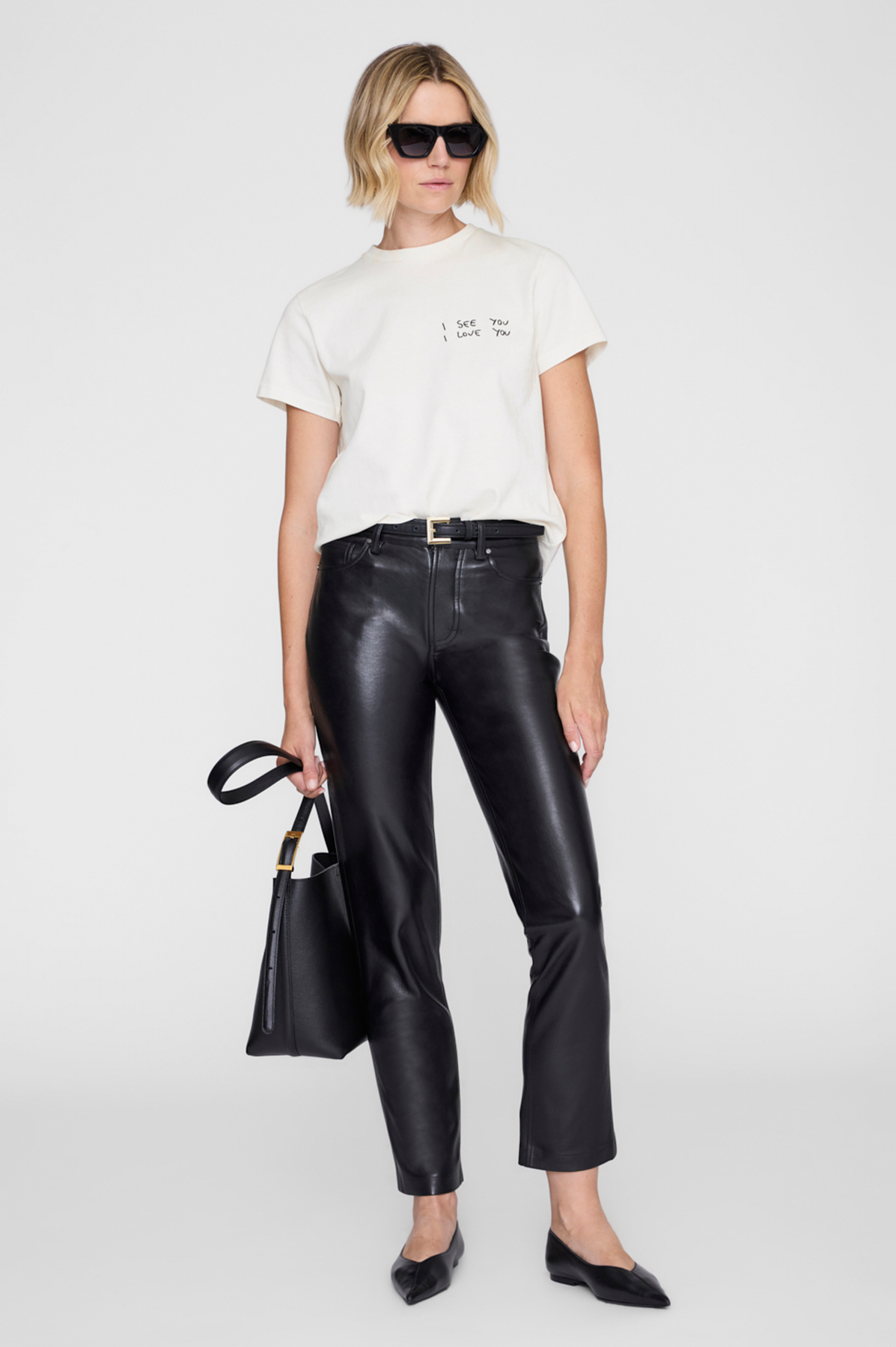 Woman wearing a white t-shirt, black leather pants, and holding a black handbag on a white background