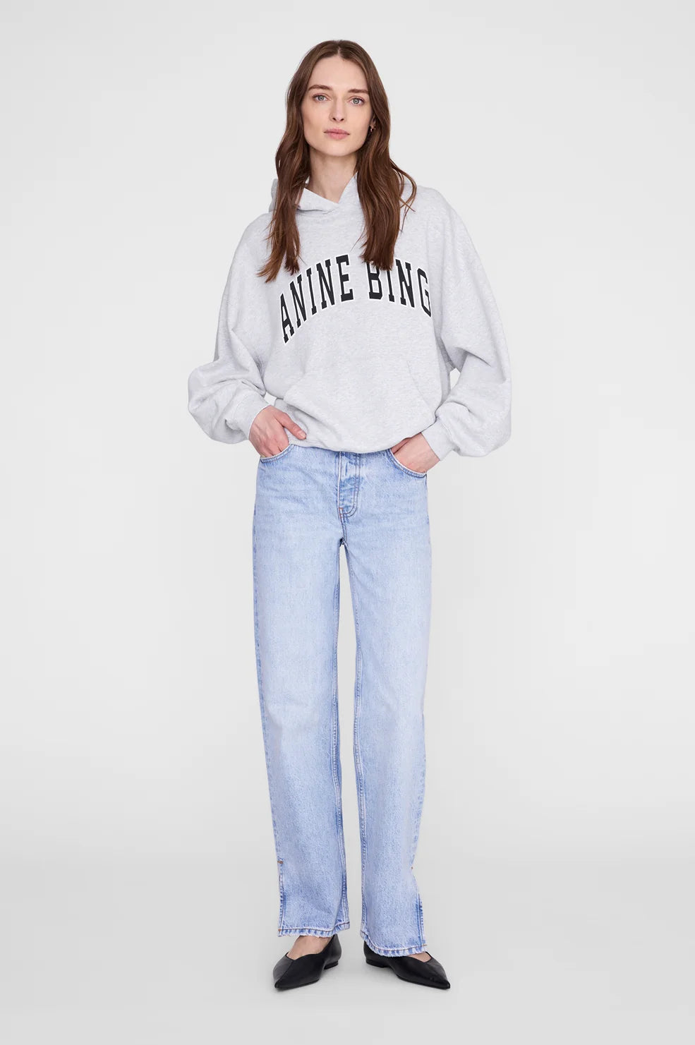 A woman modeling a light gray hoodie with 'ANINE BING' printed in black arched text across the chest, paired with light-wash blue jeans and black flats, against a plain light gray studio background.