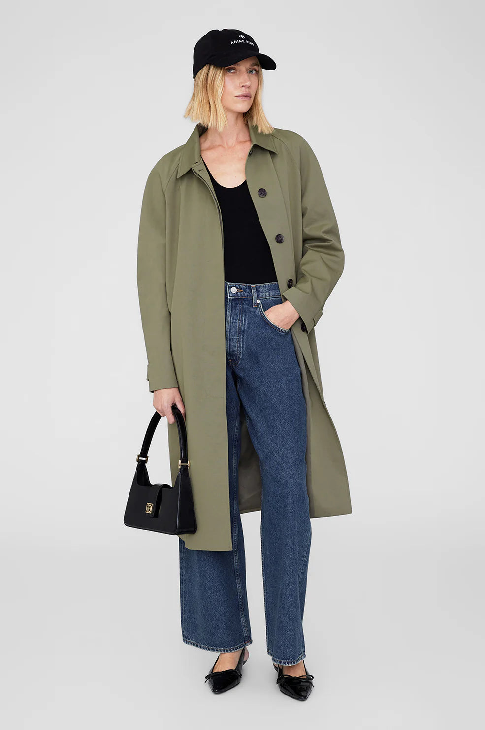 Woman wearing a green coat, black cap, black top, blue jeans, and black shoes from Anine Bing on a white background