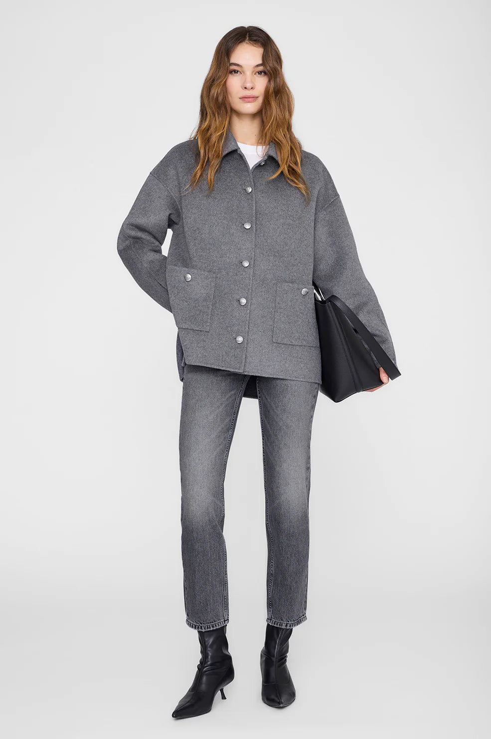 Woman wearing a gray wool jacket from Anine Bing, straight jeans, and black boots holding a black bag on a white background