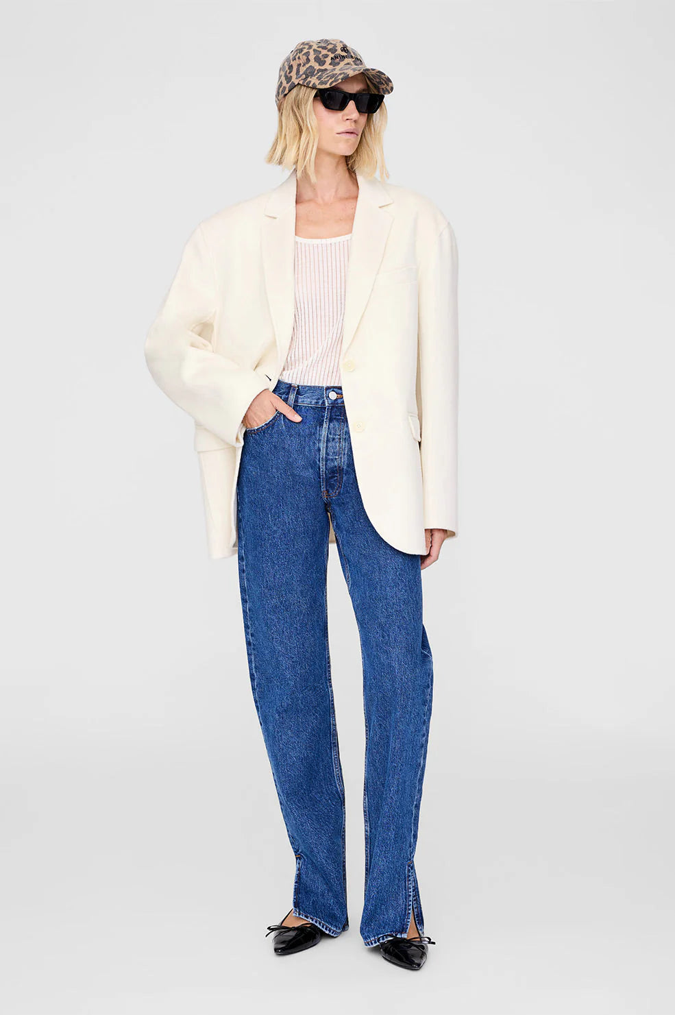 Person wearing a white blazer, blue jeans, and a leo print cap on a white background