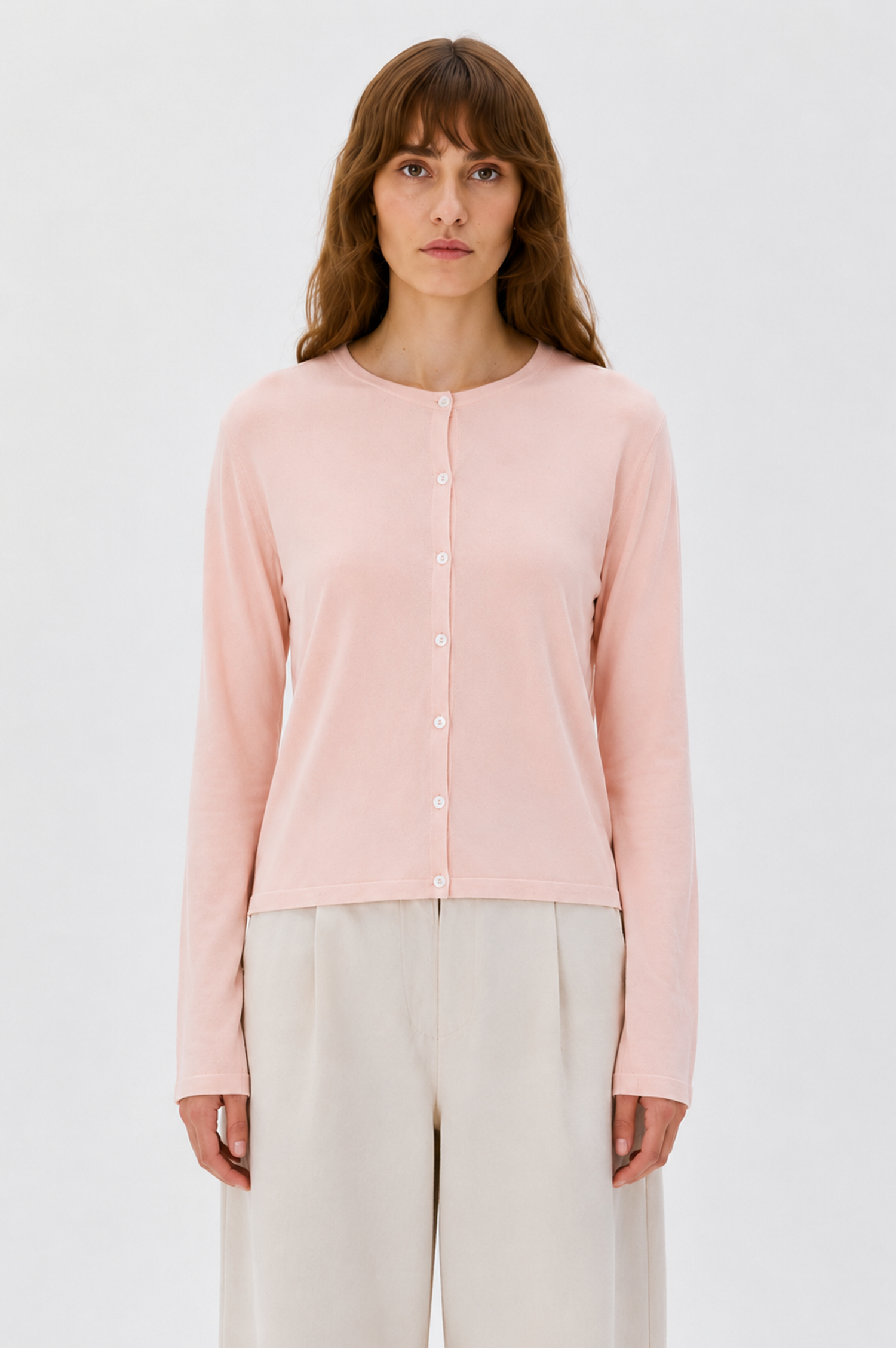A woman stands against a light gray studio background wearing a pale pink long-sleeve button-front cardigan and off-white wide-leg pants.