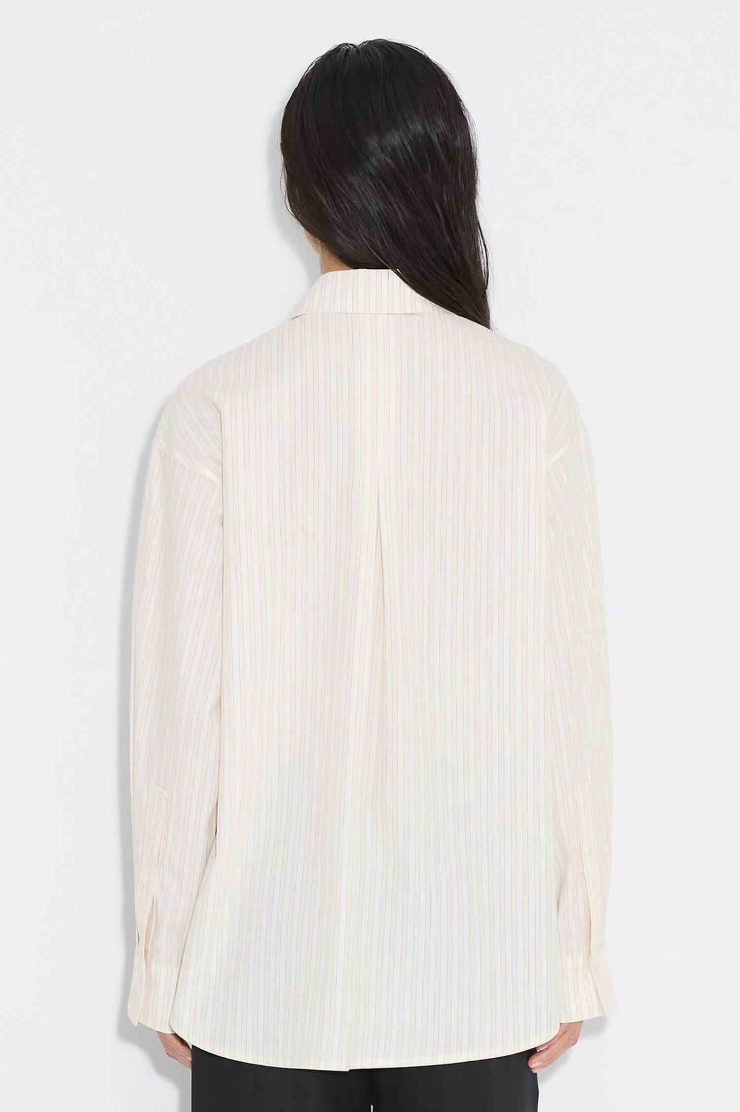 A woman viewed from the back wearing a cream-colored, long-sleeve button-up shirt with thin vertical stripes, paired with black pants, against a plain white studio background.