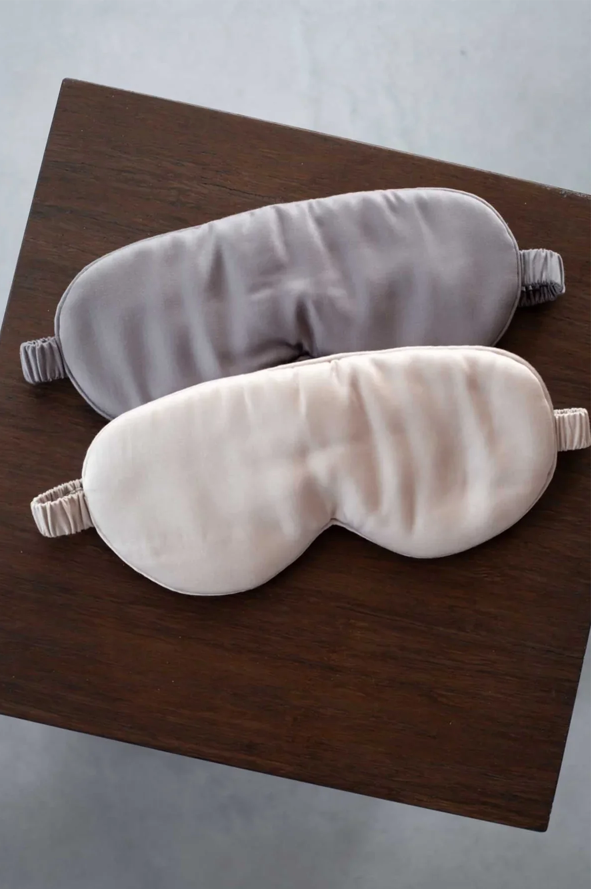 Two sleep masks, made of bamboo slk one gray and one sand on a wooden surface.