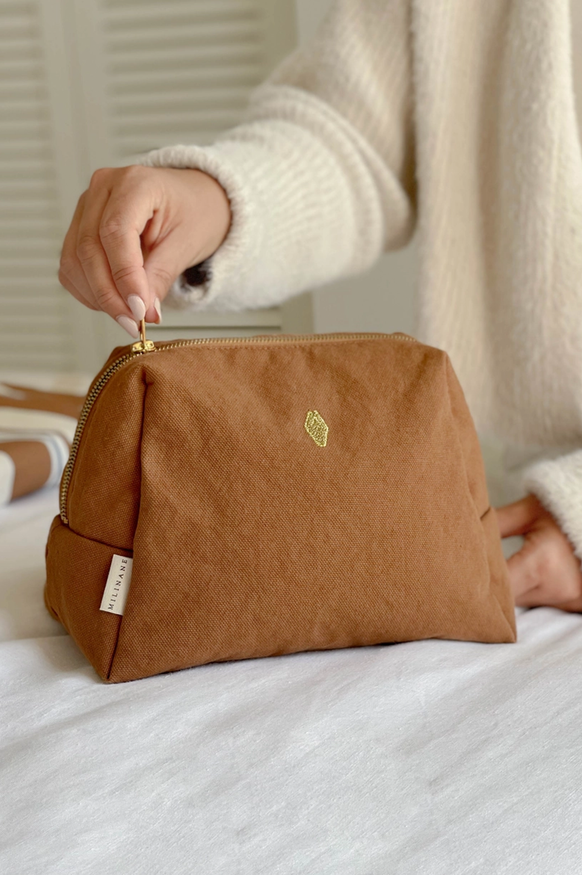 Brown toiletry bag with a gold zipper held by a person wearing a white sweater.