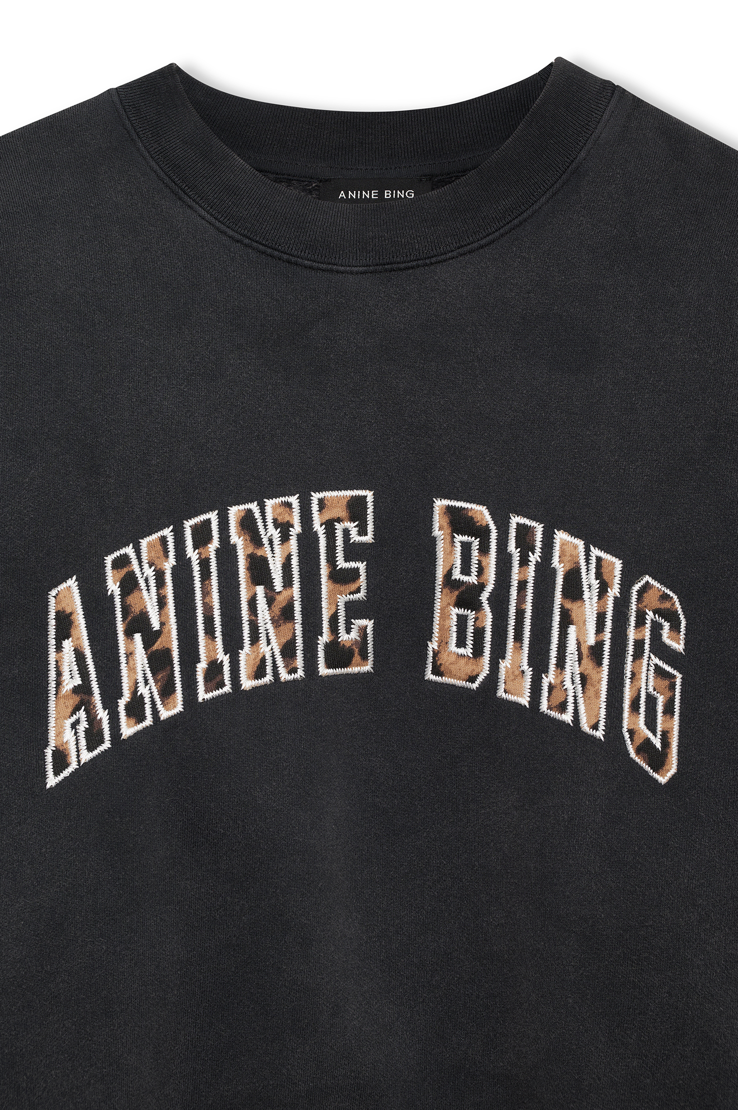 Close-up of a black crewneck sweatshirt featuring the brand name 'ANINE BING' arched across the chest in a leopard-print pattern with a white outline.