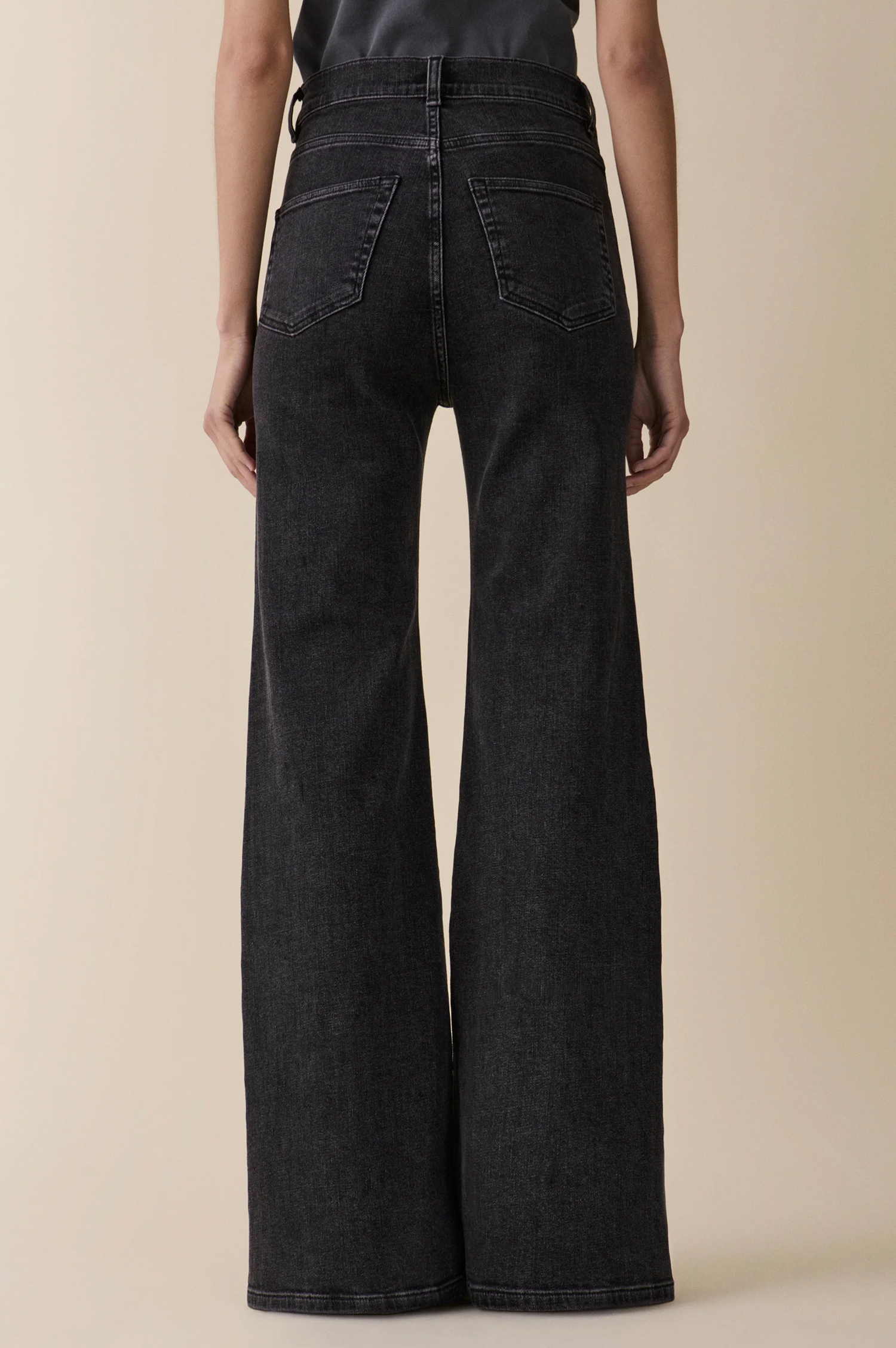 Rear view of a model wearing high-waisted, wide-leg black denim jeans from jeanerica against a solid beige background.