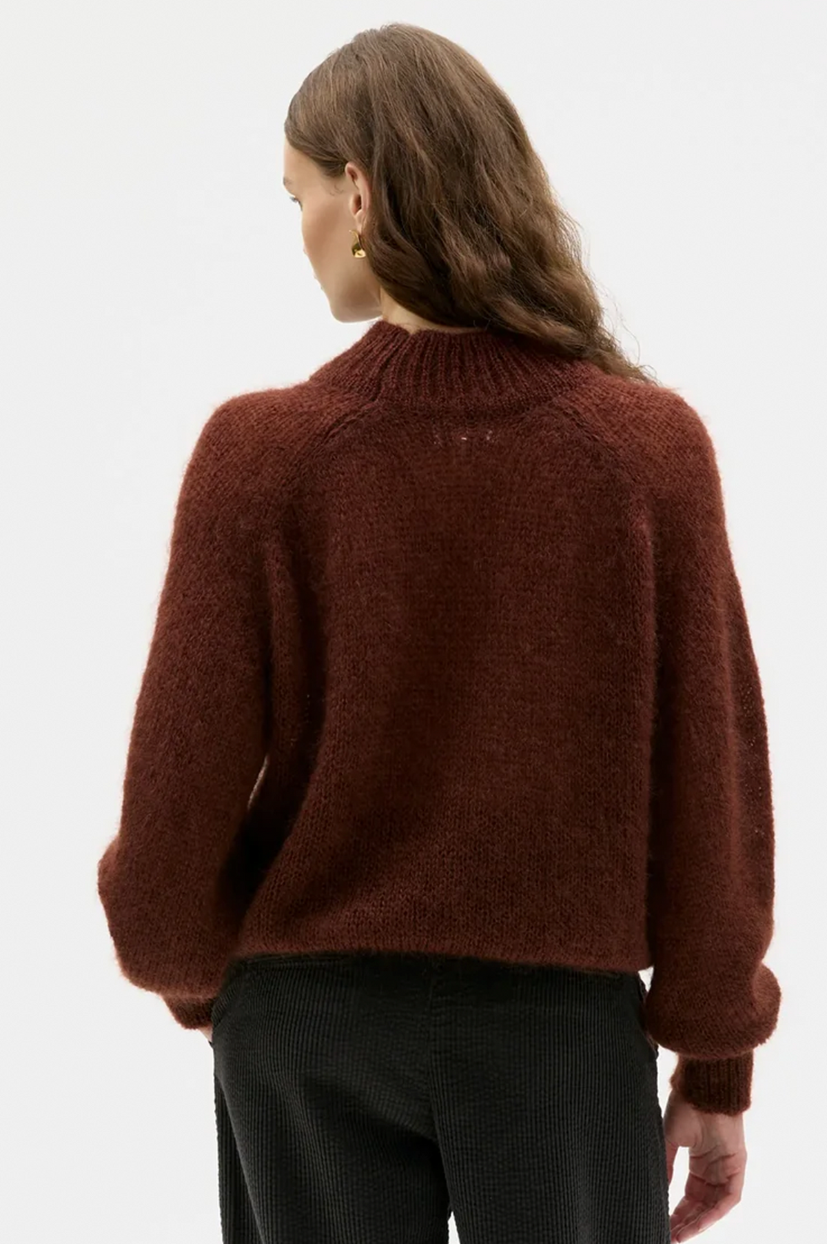A woman shown from the back wearing a fuzzy, cropped brown knit sweater and black pants against a plain white studio background.