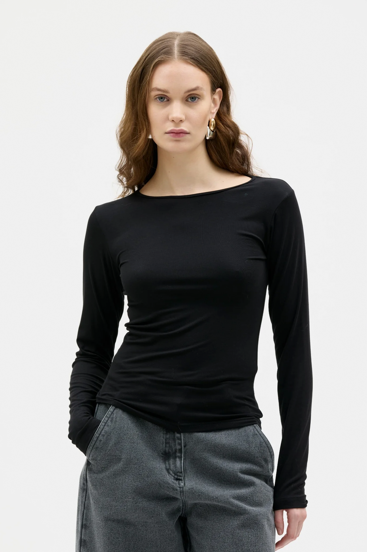 A woman modeling a fitted black long-sleeve boat-neck top with gray jeans against a plain light gray studio background.