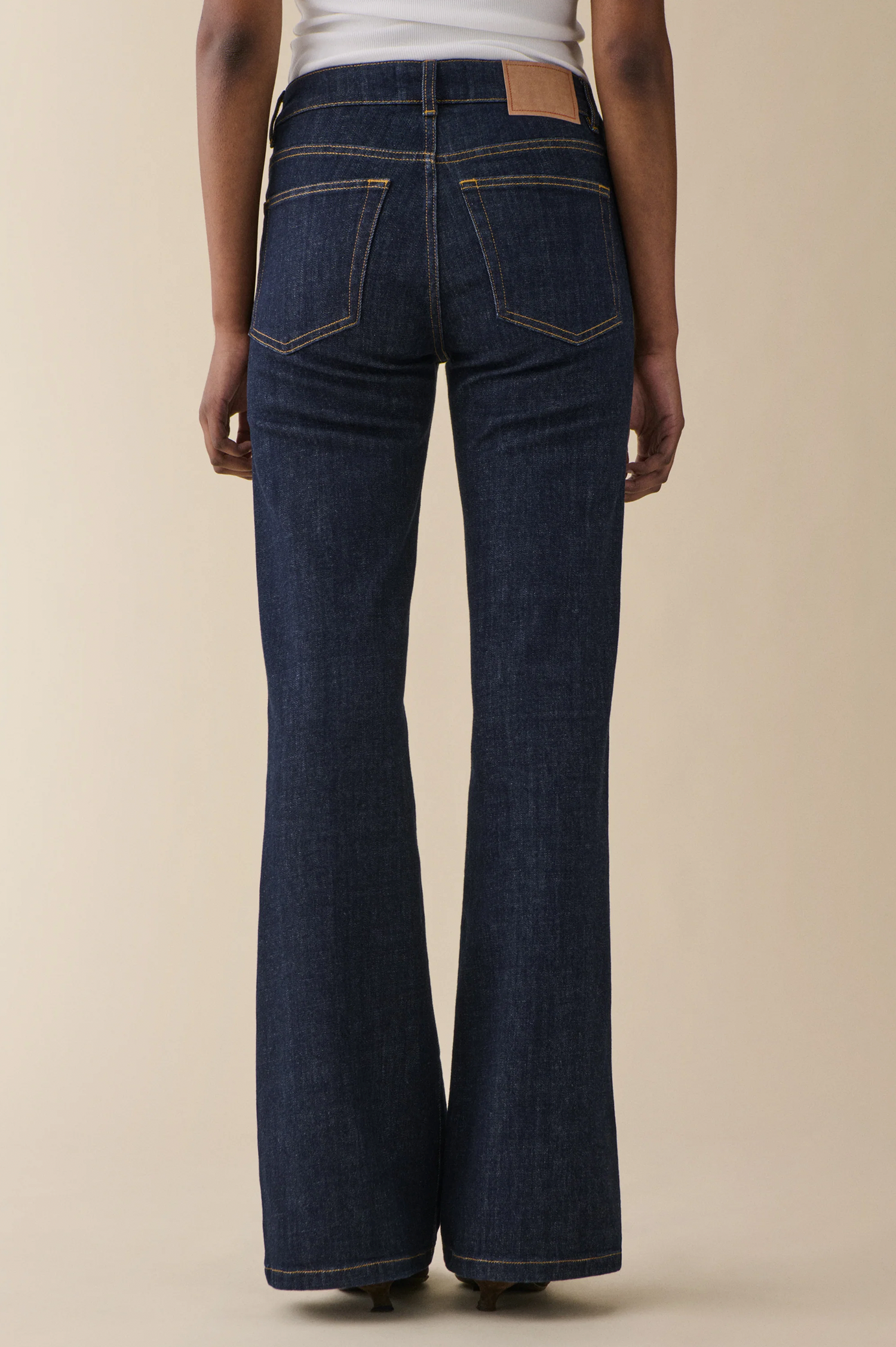 Rear view of a model wearing dark blue flared denim jeans with a tan leather patch on the back waistband, set against a solid beige studio background.