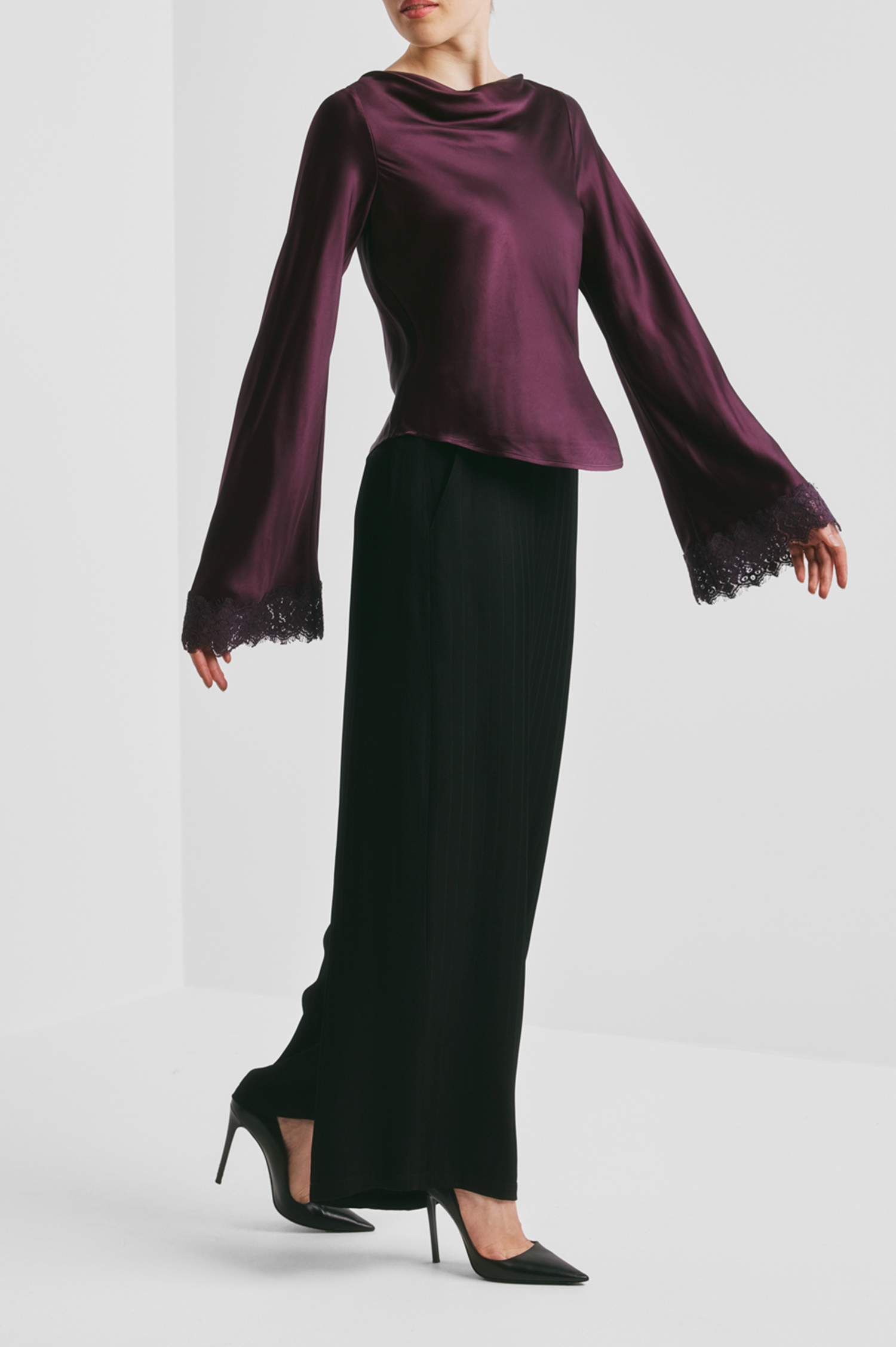 A model wearing a deep purple satin long-sleeve blouse with lace-trimmed bell sleeves and black pleated trousers, paired with black high heels, set against a minimalist light gray studio background.