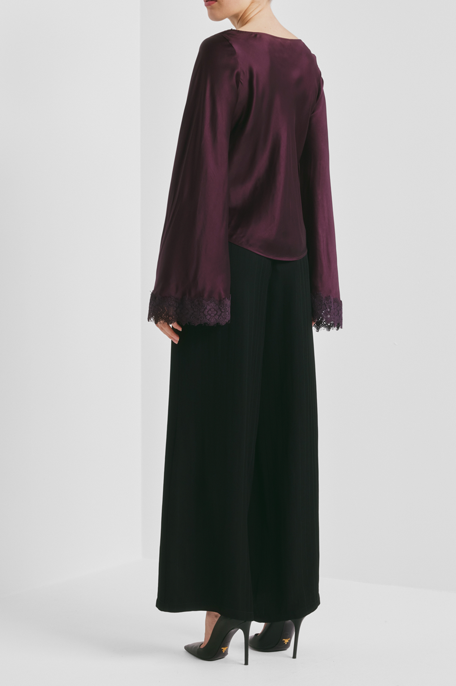 A model shown from the back wearing a deep plum purple satin long-sleeve top with lace trim at the cuffs from By Timo, paired with wide-leg black trousers and black heels.