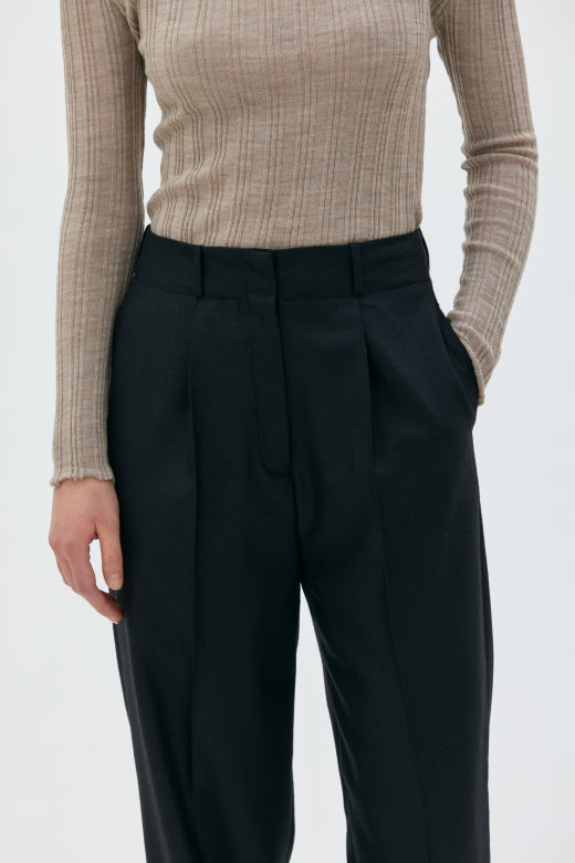 Person wearing a beige ribbed sweater and black pleated pants on a white background